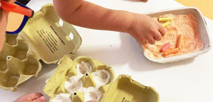 Brazo de un niños cogiendo arena y tizas de color de una caja de huevos